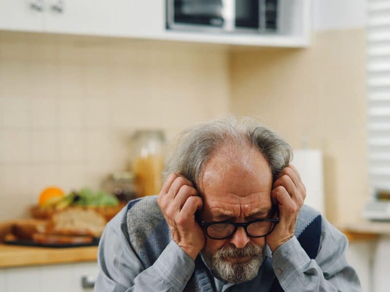 An older adult at a kitchen table reading a page of dense paperwork.