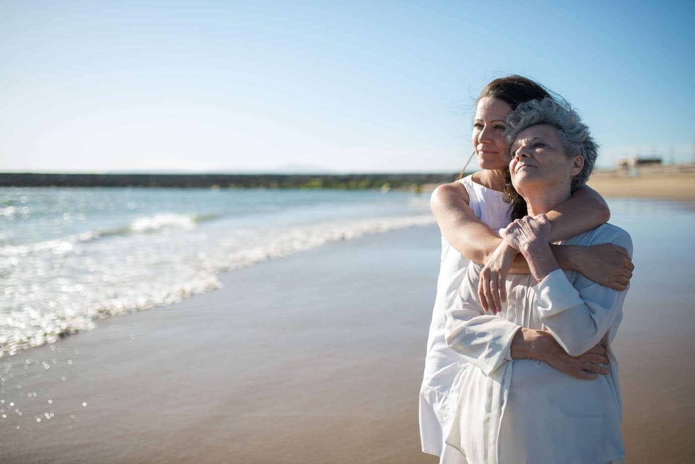 An adult daughter standing behind her elderly mother on a beach, arms wrapped around her, both looking out at the water.
